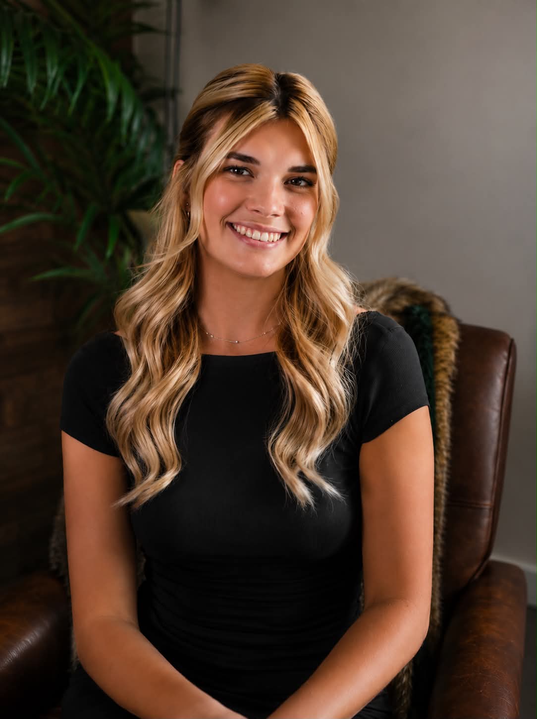 Smiling woman with long blonde hair in a black top, seated in a brown leather chair, indoors.